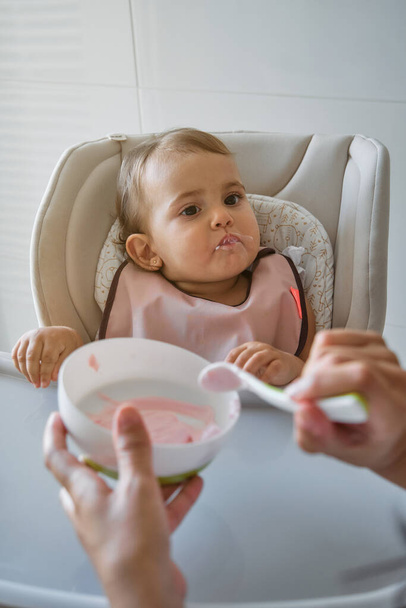 Curious baby girl in pink bib sitting on high chair and looking away while eating yogurt with help of unrecognizable person in kitchen at home - Photo, Image