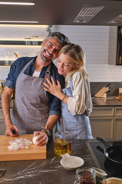Дружина обіймає позитивного чоловіка, який ріже цибулю на обробній дошці і дивиться на камеру, готуючи разом на сучасній світлій кухні - Фото, зображення