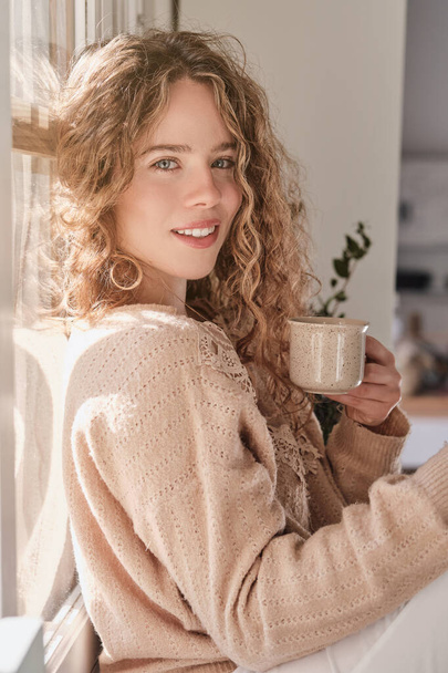 Kıvırcık sarı saçlı, pencerenin yanında çay içerken kameraya bakan genç bir kadın. - Fotoğraf, Görsel