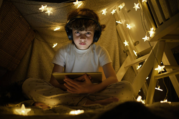 Bas angle corps complet de garçon en pyjama et écouteurs sans fil regarder la vidéo sur tablette assis dans la tente de jeu décoré avec guirlande lumineuse - Photo, image