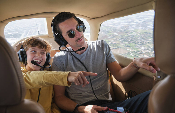 Возбужденный маленький мальчик улыбается в повседневной одежде и гарнитуре, улыбаясь и указывая в сторону, сидя в салоне самолета с молодым отцом во время экскурсии - Фото, изображение