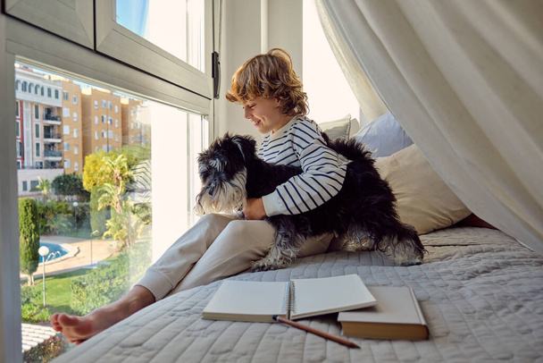 Blij schoolkind met gekruiste benen interactie met Miniatuur Schnauzer op bed met leerboek en notitieblok thuis - Foto, afbeelding