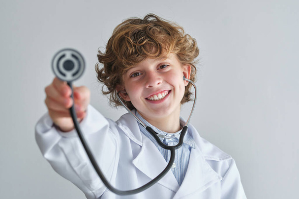 Candid παιδί σε ιατρική ρόμπα με επαγγελματικό όργανο κοιτάζοντας κάμερα σε λευκό φόντο - Φωτογραφία, εικόνα