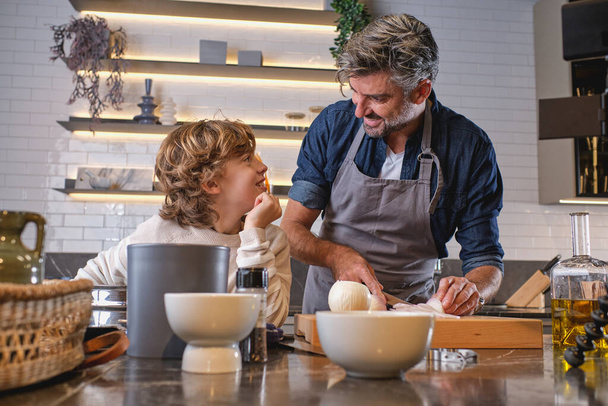Optimistic man and son looking at each other while cutting fresh onion at table with various dishware in light kitchen - Photo, Image