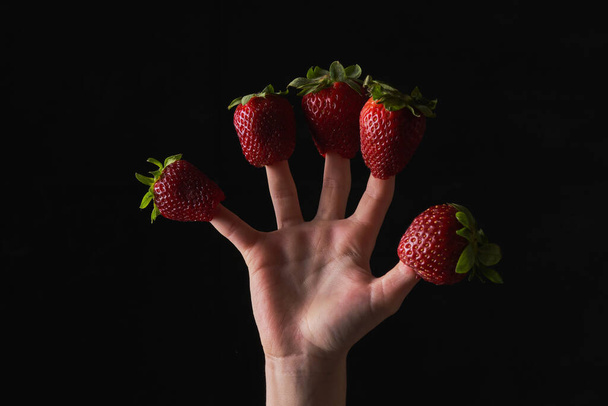 Crop unrecognizable person wearing appetizing red strawberries on fingers on black background in studio - Photo, Image