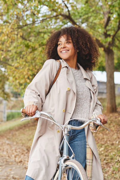 秋の公園で自転車で立ちながら笑顔と離れて見てカジュアルな服で巻き毛を持つ民族女性 - 写真・画像