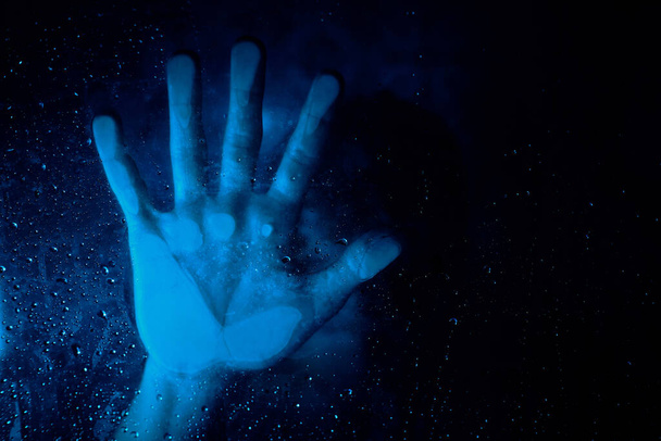 Nierozpoznawalna samotna osoba dotykająca szklanej ściany czystymi kroplami wody w niebieskim świetle - Zdjęcie, obraz