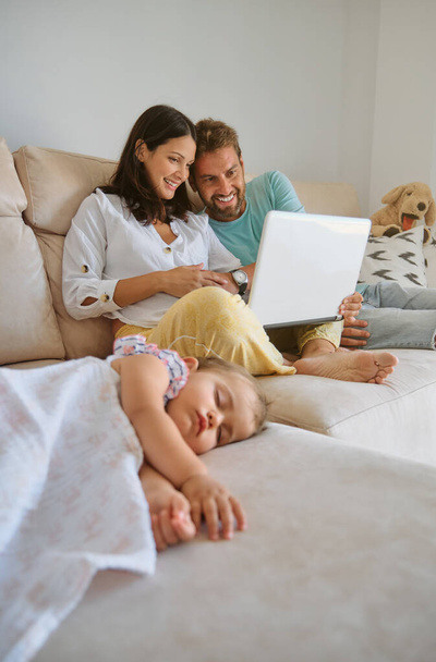 Uśmiechnięta kobieta i mężczyzna oglądają film na laptopie i siedzą na kanapie obok śpiącej dziewczynki podczas weekendowego dnia spędzonego razem w salonie w domu - Zdjęcie, obraz