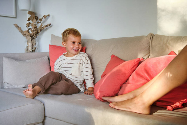 boso chłopiec w casual ubrania zabawy podczas odpoczynku na kanapie i patrząc na kobiety w salonie w domu w weekend dzień - Zdjęcie, obraz