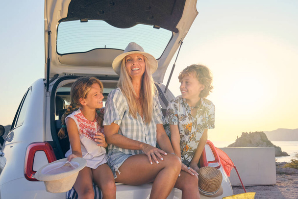 Μητέρα με δύο παιδιά με ψάθινα καπέλα και καλοκαιρινά ρούχα που κάθεται στο πορτ-μπαγκάζ ενός λευκού αυτοκινήτου κατά την ανατολή του ηλίου με τη θάλασσα στο βάθος - Φωτογραφία, εικόνα