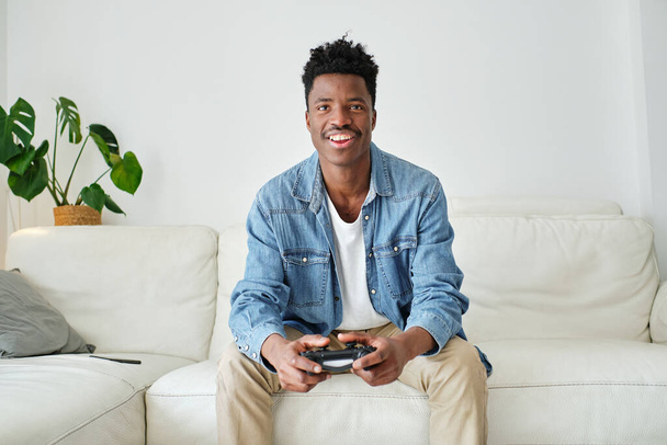 Веселий молодий афроамериканський чоловічий геймер у повсякденному одязі грає у відеоігри, сидячи на зручному дивані з джойстиком - Фото, зображення