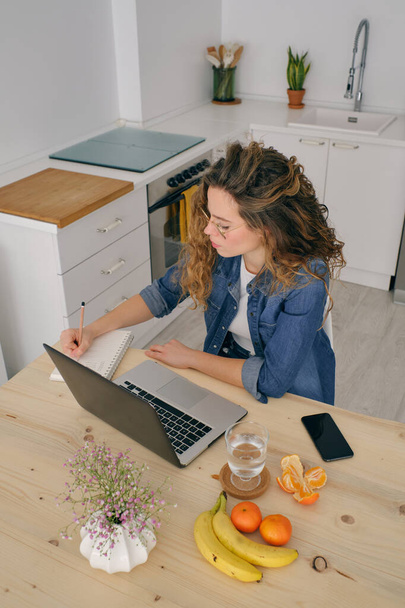 Concentrated businesswoman with long curly hair writing in notepad while working on remote project at table with netbook in kitchen at home - Photo, Image