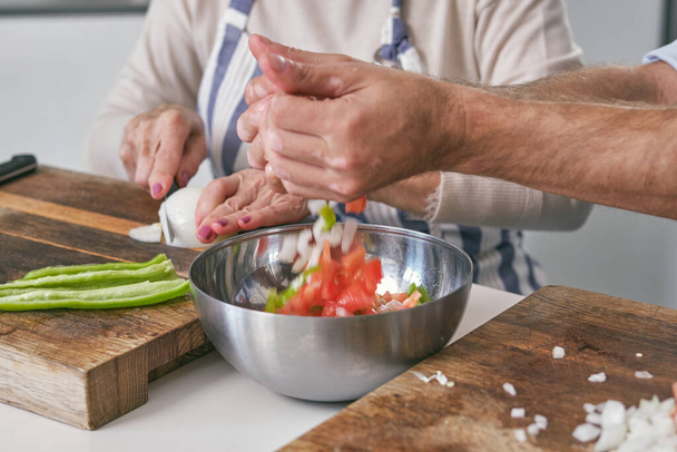 Анонимный самец, наливающий нарезанные овощи в миску, которую кладут на стол во время приготовления пищи рядом с женщиной в светлой кухне дома - Фото, изображение