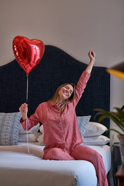 Happy female in sleepwear sitting on bed with red heart shaped balloon and raising hand while celebrating birthday and rejoicing over surprise - Photo, Image