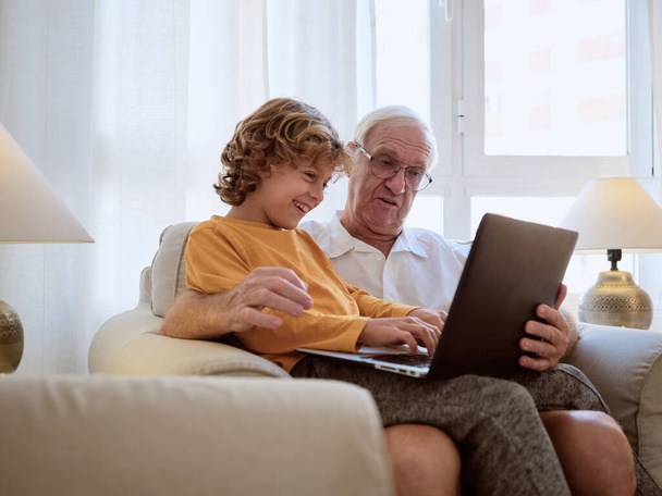 Задоволений літній чоловік з онуком друкує на сучасному ноутбуці, сидячи разом у кріслі у світлій вітальні вдома
 - Фото, зображення