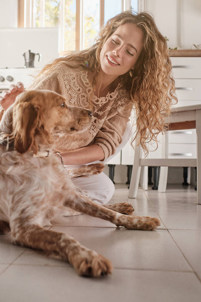 Jovem mulher positiva com longos cabelos cacheados acariciando adorável cão Spaniel enquanto sentado no chão na cozinha leve em casa - Foto, Imagem