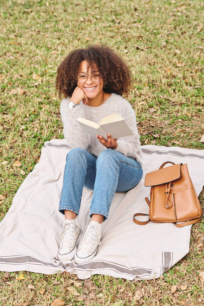 Corps complet de femme afro-américaine heureuse en vêtements décontractés souriant et regardant la caméra tout en se reposant sur la couverture et la lecture roman intéressant dans un parc vert - Photo, image