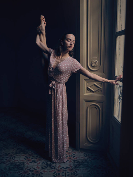 Teljes test balerina hosszú ruha áll ajtóval szemben a sötét szobában, miközben tartja a lábát, és csukott szemmel - Fotó, kép