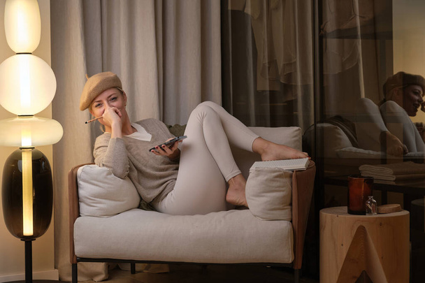 Positive woman with blond hair in beret sitting in armchair with legs bent and using mobile phone while looking at camera - Photo, Image