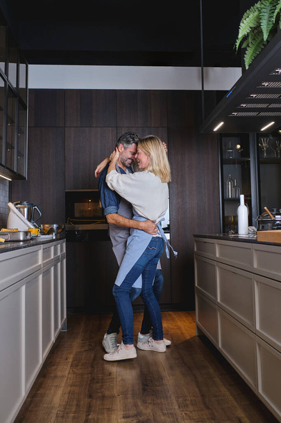 Πλευρική άποψη πλήρες σώμα του ρομαντικού ζευγαριού σε ποδιές αγκαλιάζει και χορό, ενώ περνούν το χρόνο τους μαζί στην εγχώρια κουζίνα - Φωτογραφία, εικόνα