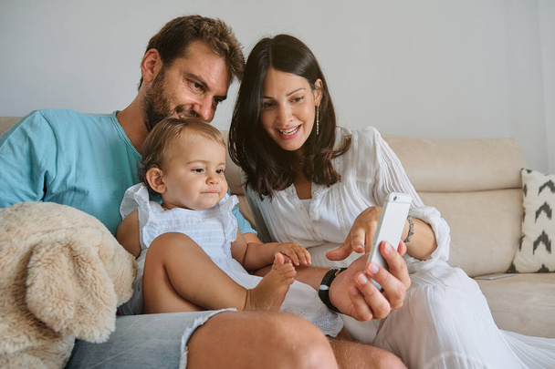 Счастливые родители с дочкой сидят на диване и смотрят видео по мобильному телефону в выходной день дома - Фото, изображение