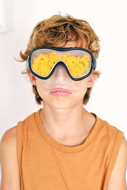 Child with brown hair in undershirt and diving goggles with yellow seaweed on light background - Photo, Image