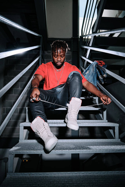 Full body of young African American male in trendy outfit sitting on stairs and tying shoelaces on fashionable sneakers - Foto, immagini