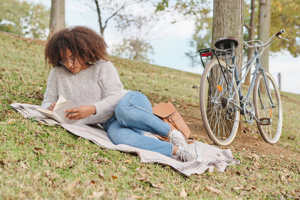 Concentrated ethnic woman in eyeglasses resting on blanket near bike and reading interesting novel in green park on weekend day - Photo, Image