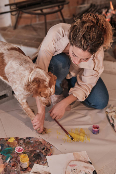 Creative female artist bonding with pet using paintbrush and paw print while drawing on fabric in modern studio - Photo, Image