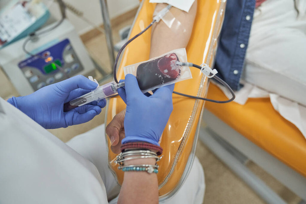 Hohe Winkel der nicht wiederzuerkennenden Krankenschwester in medizinischem Mantel und Handschuhen Kontrolltasche während der Durchführung von Blutspende Verfahren an einer Patientin in zeitgenössischen Krankenhaus - Foto, Bild