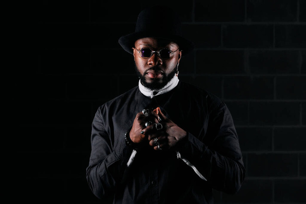Modelo masculino afroamericano seguro en traje negro con accesorios de pie contra la pared de ladrillo en habitación oscura y mirando a la cámara - Foto, Imagen