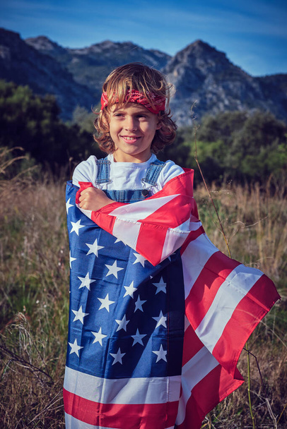 Onnellinen söpö esiteini poika seisoo ruohoisella niityllä käärittynä Amerikan yhdysvaltojen lippuun - Valokuva, kuva