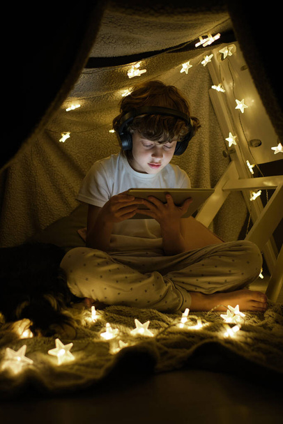 Corpo inteiro de menino sério em fones de ouvido assistindo vídeo no tablet moderno enquanto sentado com pernas cruzadas perto de guirlanda brilhante no quarto escuro - Foto, Imagem