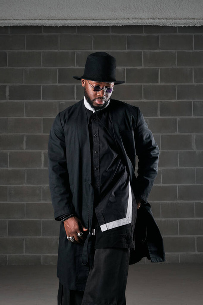 Serious bearded black male in trendy outfit and hat standing near brick wall with hand in pocket and looking down - Photo, Image