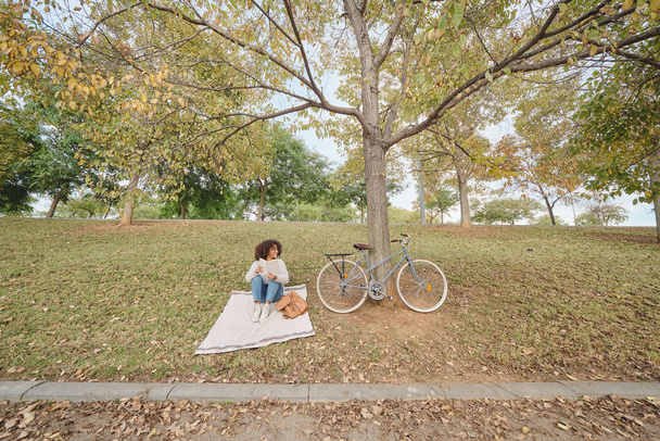 Femme afro-américaine en vêtements décontractés reposant sur une couverture sur l'herbe et lisant un livre intéressant dans la nature le week-end - Photo, image