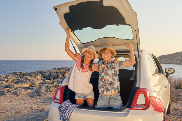 Twee kinderen buigen in de kofferbak van de auto met hun handen omhoog in een gebaar van overwinning terwijl ze naar de camera kijken en dragen een strohoed met manden van voorwerpen voor het strand naast de auto met de zee op de achtergrond - Foto, afbeelding
