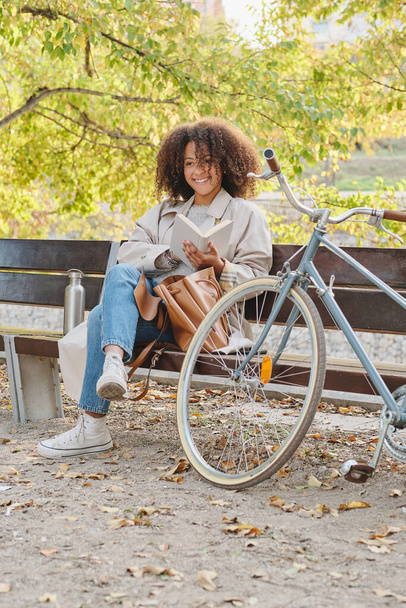 Cuerpo completo de joven estudiante étnica positiva con pelo afro descansando con libro en banco de madera en el parque cerca de la bicicleta - Foto, Imagen