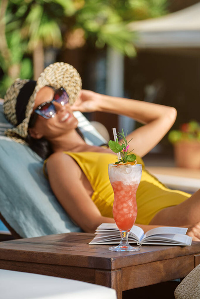Seitenansicht einer jungen Frau in Badeanzug und Sonnenbrille, die neben einem Tisch mit einem Glas frischem Getränk ruht - Foto, Bild