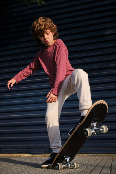 Full length boy doing skateboard trick on pavement against dark blue wall on sunny day in city - Foto, afbeelding