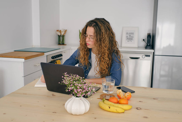 Koncentrált női vállalkozó alkalmi ruhák ül fa asztal gyümölcsökkel és jegyzeteket jegyzetel, miközben böngészi netbook és dolgozik a távoli projekt konyha otthon - Fotó, kép