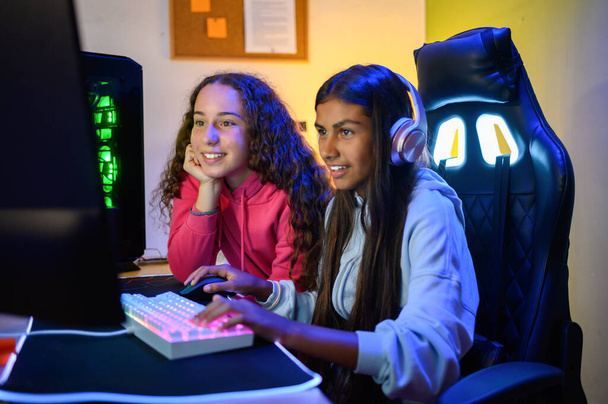 Positieve tiener multiraciale kijken naar het scherm van de monitor tijdens het zitten aan tafel met gaming PC en met behulp van toetsenbord om spel te spelen in de kamer - Foto, afbeelding