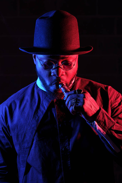 Etninen parrakas mies mustassa hatussa tupakointi hookah seisoessaan punaisella ja sinisellä neonvalolla tummaa taustaa vasten - Valokuva, kuva
