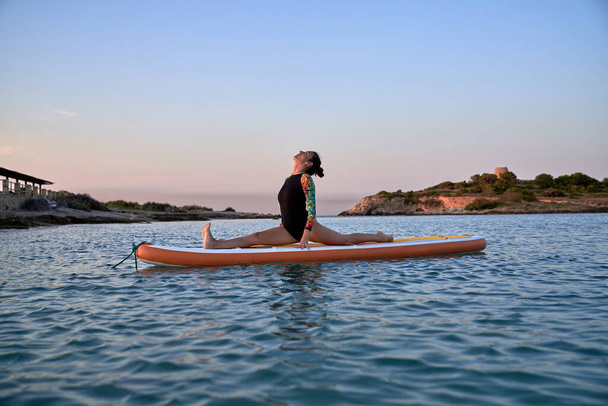 Vista lateral da jovem em trajes de banho tomando postura Hanumasana enquanto pratica ioga no paddleboard à beira-mar à noite - Foto, Imagem