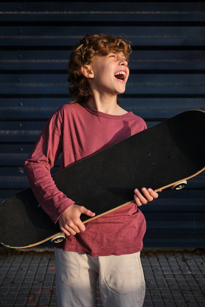Piccolo pattinatore felice con skateboard ridere e distogliere lo sguardo mentre si riposa sulla strada della città - Foto, immagini