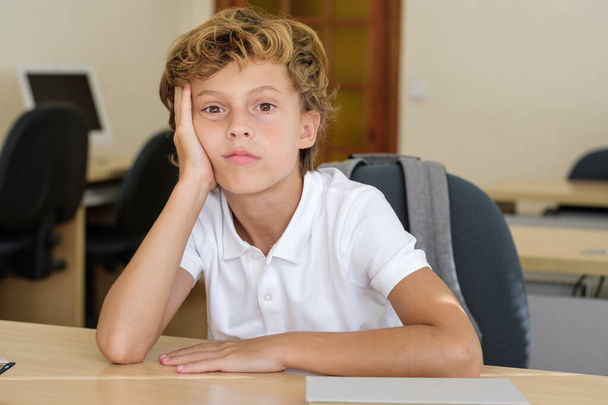 Bored schoolkid leaning on hand at wooden desk in classroom and looking at camera - Photo, Image