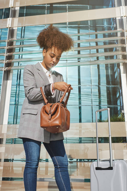 Düşük açılı Afro-Amerikan bayan gezgin şık giyimli ve bagajı şehrin cam havaalanı duvarının yanında duruyor. - Fotoğraf, Görsel