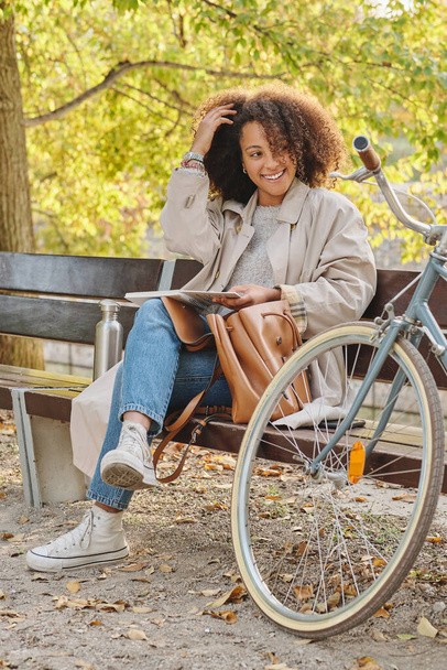 Celé tělo mladé pozitivní stylové etnické dámy odpočívající s knihou na dřevěné lavičce u kola v parku a dotýkající se vlasů - Fotografie, Obrázek