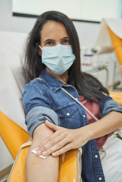 Γυναίκα με μαύρα μακριά μαλλιά στη μάσκα βάζοντας λευκό βαμβάκι στο χέρι μετά από να δώσει αίμα, ενώ κάθεται σε πορτοκαλί καρέκλα στο κέντρο αιμοδοσίας - Φωτογραφία, εικόνα