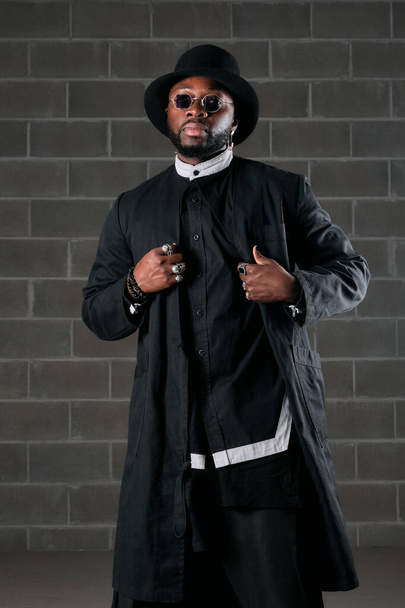 Уверенный афроамериканец в модном наряде и солнцезащитных очках, стоящий у кирпичной стены и смотрящий в камеру во время прикосновения пальто - Фото, изображение