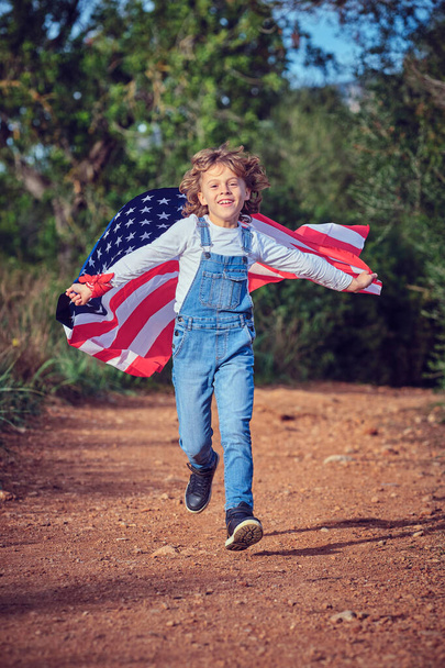 Повне тіло чудового хлопчика дивиться на камеру під час бігу по дорозі з національним махаючим американським прапором в літній день - Фото, зображення